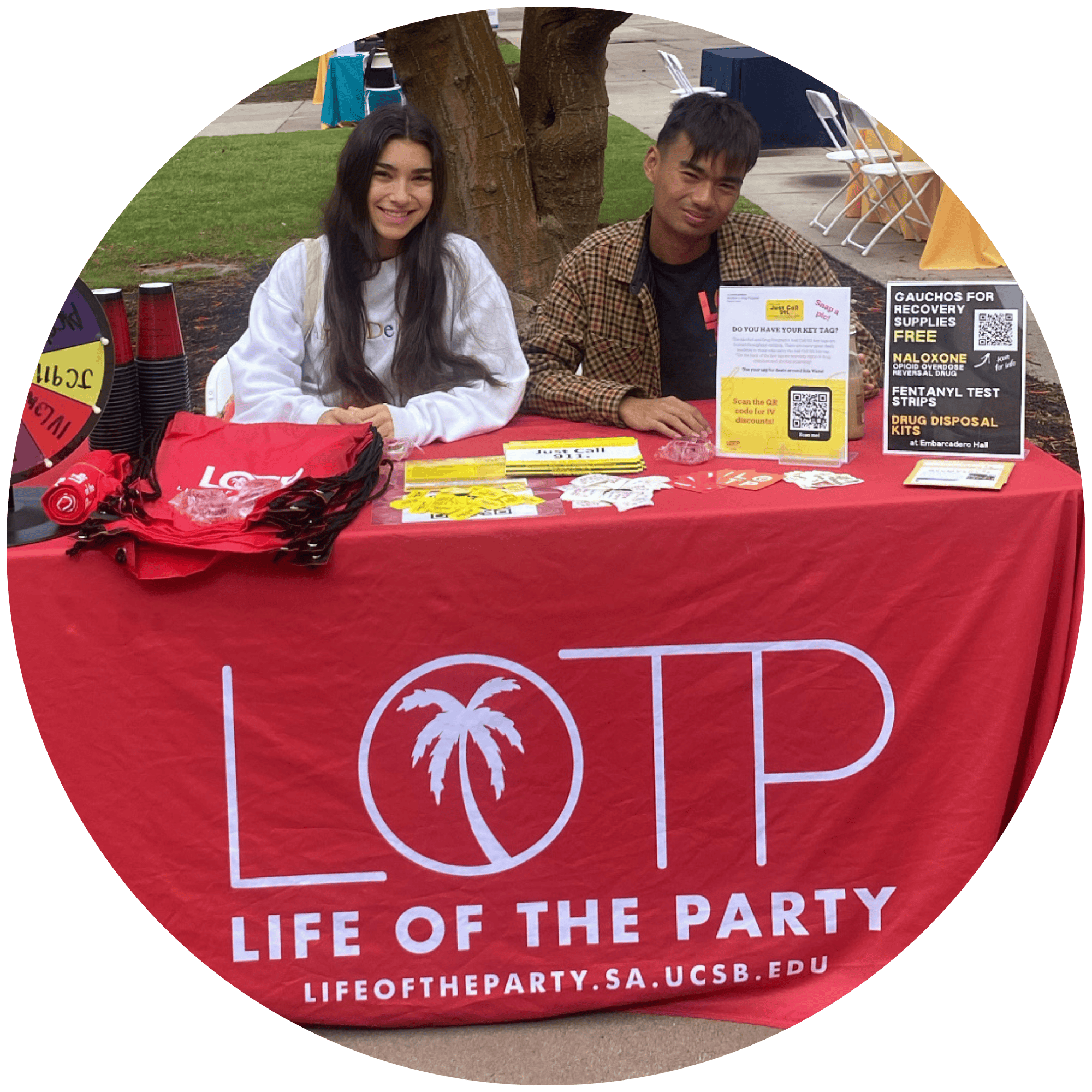The image depicts two members of the Life of the Party (LOTP) team sitting at a table with a red tablecloth that reads LOTP.